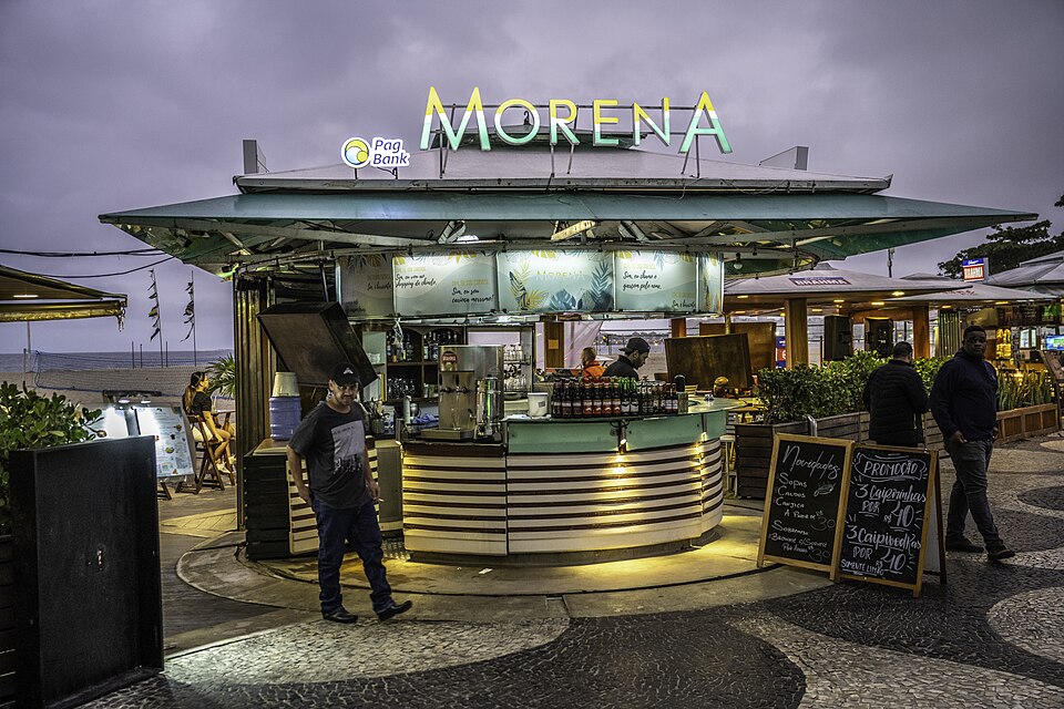 A lit beach kiosk on the wave-patterned promenade at dusk